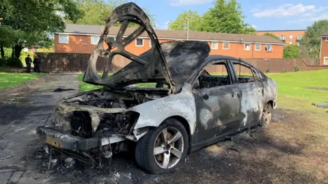 A car which has been burnt out and and is completely destroyed is on the grass on a residential street.