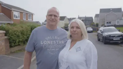 Chris and Jane Williams standing on their residential street. Chris has white hair and glasses and is wearing a light blue t shirt, while Jane has bobbed white hair and is wearing a white blouse with blue diamonds on it. In the background you can see houses and cars.