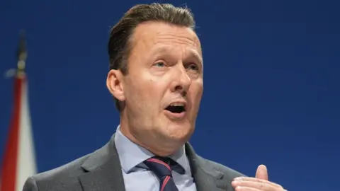 PA Media Russell Findlay, who has short dark hair, speaks at podium in front of a blue background. He is wearing a grey suit, blue shirt and blue and pink tie. 