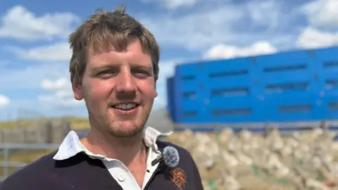 BBC Aaron with short brown hair smiling at camera. He is wearing a navy jumper with a white collar. He has a small microphone with a grey fury wind shield on his collar. The background is blurred and has sheep and a blue transport lorry