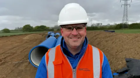 John Fairhall/BBC A head and shoulders image of a man wearing a white hard hat and an orange high-vis jacket. He is smiling and looking into the camera while standing in a field amongst piles of dirt.