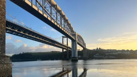Tamar bridge spanning a river at sunset, with reflections in calm water and houses on the far bank.
