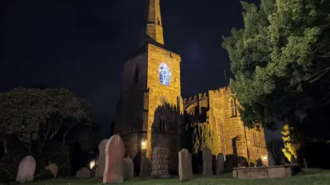 An illuminated church, shown at nighttime, surrounded by trees and a graveyard. There is a square tower with a conical spire and a large adjoining building with stained glass windows.