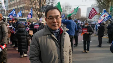 A middle-aged Korean man wearing a thick coat is smiling at the camera, while in the background are protesters holding protest flags and South Korean and American flags