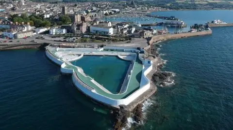 An aerial view of Jubilee Pool in Penzance. The pool is triangular shaped with the sea surrounding it. There are houses in the distance. 