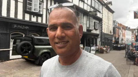 Becca Simmonds/BBC A man with short dark hair smiles at the camera on a town centre street, backed by black-and-white half-timbered buildings. He has glasses on top of his head and is wearing a grey T-shirt. 