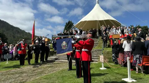 Trumpeters in red uniforms play in front of the hill, which is covered by a canopy, while dignitaries look on.