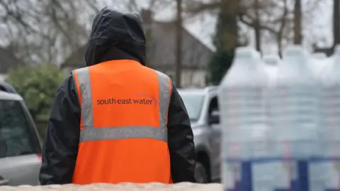 A person in a black coat and orange vest which says "South East Water" on the back. There are water bottles stacked up to the right of the person, and two silver cars in the background