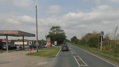 Google A single-carriageway road with a garage forecourt on the left, which has been converted into a car wash. There are trees and a grass verge on either side of the road.