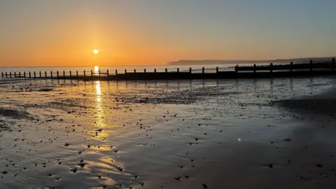 The sun rising at Redcar beach. The sky is tinted orange and the tide is out, with the sunlight glowing on the wet sand in the foreground. The silhouette of a headland is visible in the distance. 