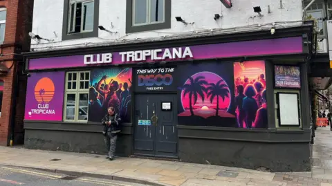 A man stands outside the purple and pink frontage of Club Tropicana. 