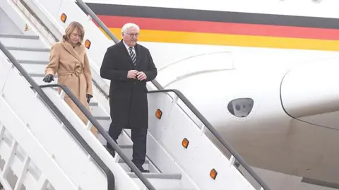 Getty Images A man and a woman walking down the steps of a plane liveried in the German flag. She has fair hair and is wearing a long beige coat; he is in a long black coat, white shirt and black tie and has white hair.