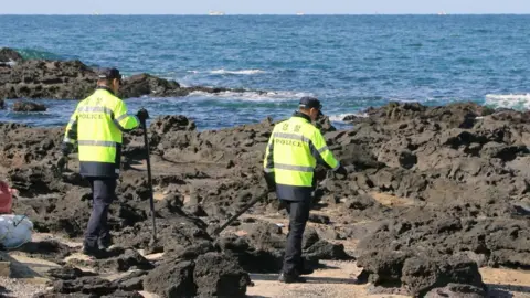 News1 Two police officers, in flourescent yellow jackets, use sticks to comb through the rocky shore