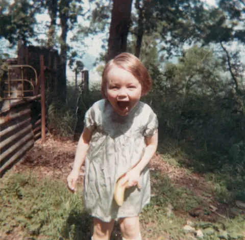 Supplied A young girl with short red hair and a dress, holding a banana. She is standing outside in a field with a gate to the side