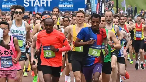 A large group of runners crowd the road at the start of a marathon, running closely together beneath a blue “START” banner marked “Belfast.” In the foreground, several runners wear brightly coloured singlets and visible race bibs, including numbers 23 and 24, while many others follow behind. The scene shows a busy, competitive race atmosphere with runners filling the width of the road.