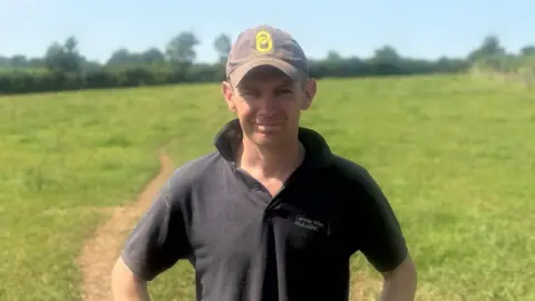 Rob wears a dark green polo shirt and a baseball cap and has his hands on his hips. He is standing in a grassy field with hedgerows and trees in the distance. 