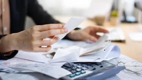 Getty Images A close-up of someone looking at household bills with a calculator in front of them on a desk next to a pair of glasses