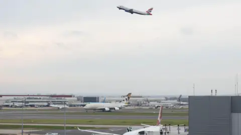 A view of Heathrow airport - several planes are sitting on runways, while one plane - a British Airways plane with a Union Jack design on the tail - is going into the sky.