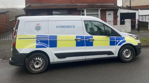 A white van is parked outside a house. It has yellow and blue police livery and the word 'forensics' on the side of it alongside the Staffordshire Police emblem.