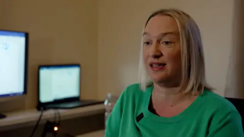 Anna Bruce-Hou, who has blonde shoulder-length hair and wearing a green jumper, is sat in a room with blank walls. Two computer screens can be seen in the background.