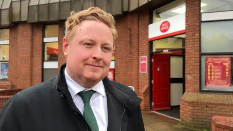 BBC A man with short blonde hair and earing a black jacket, white shirt and green tie, stands outside the entrance to a brick building with windows and a red door with the words POST OFFICE above it.