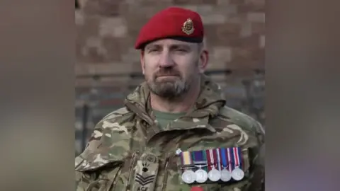 Combat Stress Rowley had a brown and grey beard and facial hair, with a green, white and brown camoflage jacket on. There are five silver medals with colourful ribbons on the breast of his jacket