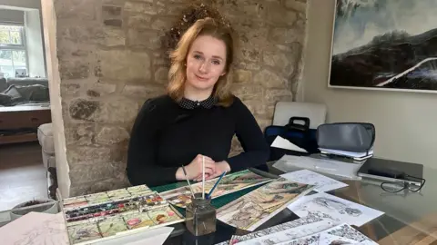 A female artist has paints and watercolour paintings on a table in front of her