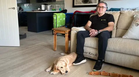 BBC A golden Labrador puppy, Chrissie, is lying on a wooden floor in front of a beige sofa. Richard is seated on the sofa, and a long brown chew toy is on the floor near the puppy. The background shows a modern kitchen with black cabinets, a green suitcase, and framed artwork on the wall.