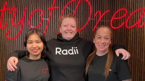 Three women wearing dark tops smiling and standing in front of a red neon sign which reads: 'City of Dreams'.