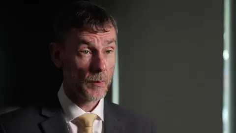 A man in a black suite and golden tie speaks in a dark room with a serious expression. 