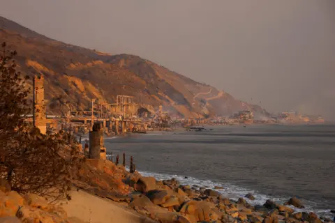 Mike Blake/Reuters Asap mengepul dari rumah-rumah tepi pantai yang terbakar di sepanjang jalan pesisir menuju Malibu pada hari Rabu, menunjukkan deretan bangunan yang hancur akibat kebakaran.