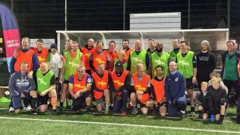 BBC A group of football players wearing high visibility bibs in orange and green stand together as a team by the dugout on the side of the pitch. Former England player Emile Heskey is in the centre.