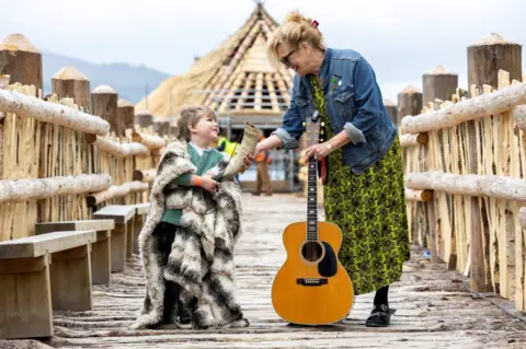 Martin Shields Barbara Dickson, a 78-year-old singer and musician with four-year-old Albie Dunlop stand in front of the recreation of an Iron Age roundhouse. 