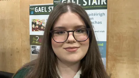 A teenage girl with long brown hair and glasses. She is wearing a school uniform.