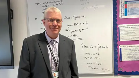 Clare Worden/BBC Head of alternative provision at NCC Andy Tovell stands in front of a white board with a series of complicated maths formula written on it. He is wearing a dark grey suit and a lilac shirt with matching patterned tie. He has short white hair and glasses. 