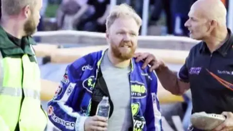 Rider Cameron Heeps walking away after the crash. His team are surrounding him. He is holding a bottle of water.