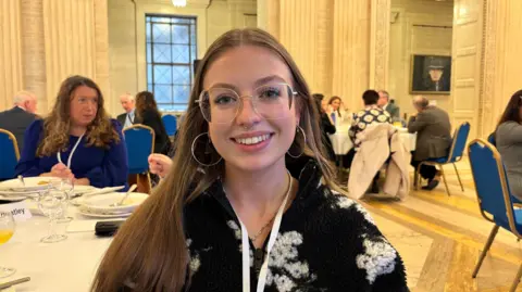 Eva Hanna is facing the camera and is smiling. She has long fair hair and is wearing glasses. In the background there are people sitting at dinning tables with plates and cutlerary.