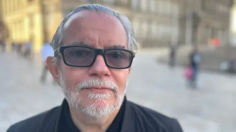 A man with grey hair and grey beard wears black glasses and a black jacket with a black top underneath.