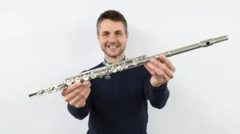 Gardiner Houlgate Auction House A man with short dark hair and a dark top holding up a silver flute