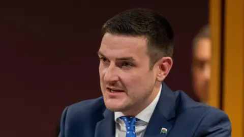 ames Evans is wearing a blue suit, blue tie and white shirt, in front of a dark background. He is looking away from the camera.