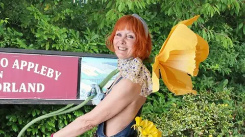 A woman with orange hair smiles at the camera. She has a large yellow flower behind her. She is wearing dungarees and a spotted neckerchief, but her arms and back are not covered. The name Appleby is written on a sign.