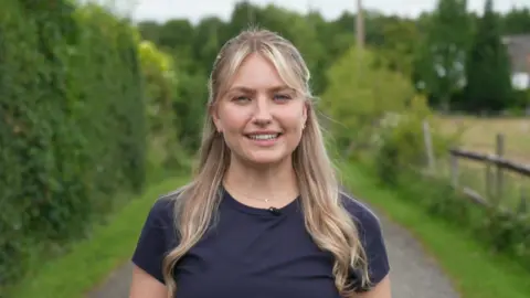 Wearing a navy blue round-necked T-shirt, Amy Thompson is standing on a countryside path surrounded by hedges and trees. Amy, with long, blonde hair, is smiling directly at the camera.