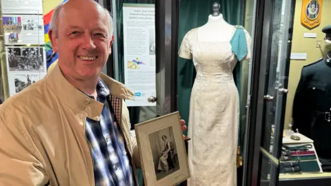 A man holding a black and white framed picture in front of a white dress