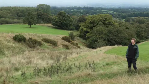 A man standing with his hands in his pocket on some land. The land slopes downwards and into the distance, with long grass in the foreground and green fields and trees in the background.