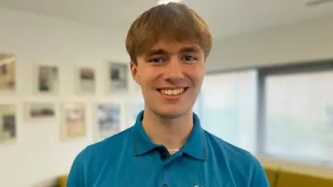 A man with short blonde hair smiles into the camera. He is wearing a blue top and is standing in an office within the Sewell Group headquarters in Hull.