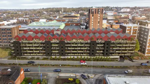 Centric&Centric Artist impression of the apartments in place of the car park. Small, red roofs sit atop the former car parks with apartments on many levels below with green indoors or outdoor plants visible from the front of the building. The building is next to a road in the town centre with taller properties surrounding it.

