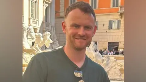 Supplied A head-and-shoulders shot of George Foster on holiday in a foreign city, possibly Rome. He is smiling and is wearing a grey T-shirt. A pair of sunglasses is hooked over the neck of the T-shirt. He has short brown hair and a short beard and moustache. Behind him, there is a civic statue and an apartment building.