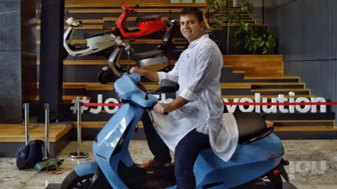 Getty Images Indian entrepreneur, co-founder and CEO of Ola, Bhavish Aggarwal, wearing a light grey Indian kurta poses for a photograph with the blue Ola electric scooter during its launch at the Ola headquarters in Bangalore on August 15, 2021. 