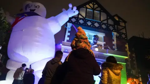 The Halloween House with the Marshmallow Man on the left and a small child on an adults shoulders looking at the hose. The child is wearing an orange hat and orange jacket. There are people either side of them also looking at the house.