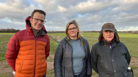 Martin Heath/BBC Simon Collyer in a red and orange anorak, Aska Pickering in a grey top and grey coat and Andrew Stevenson in a dark green raincoat, standing in a green field. There are trees and hedges along the border in the background.